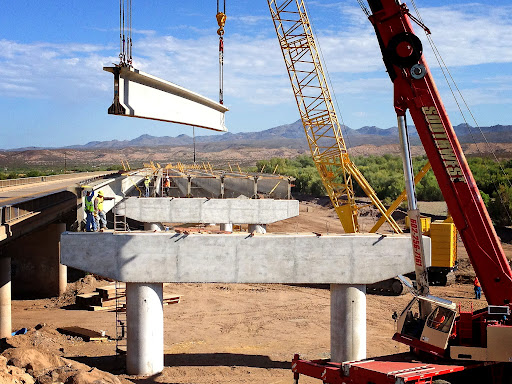 Construction workers and cranes installing large concrete beams on a bridge under construction in a desert landscape with mountains in the background.