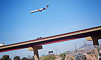 A commercial airplane flies low over a highway overpass with cars, against a clear blue sky and distant mountains in the background.