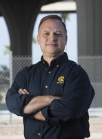 A man stands outdoors with arms crossed, wearing a black button-up shirt with a yellow logo, in front of a chain-link fence and large architectural columns.