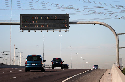 Cars driving on a highway under an electronic sign displaying travel times: “MINUTES TO SR-143: 10, I-10 TUNNEL: 15,” with a clear sky and power lines overhead.