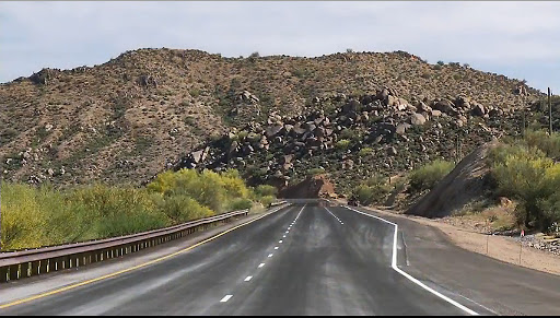A two-lane highway leads toward a rocky hill covered with sparse desert vegetation under a partly cloudy sky. There are guardrails and shoulder areas on each side.
