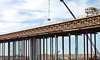 A crane lifts construction materials onto a long, elevated bridge supported by tall pillars, with a partly cloudy sky in the background.