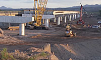 Construction equipment working on a bridge with unfinished concrete supports and dirt piles nearby.
