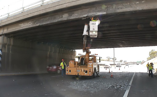 Workers use a bucket truck to inspect and repair damage under a highway overpass while debris is scattered on the closed roadway below.