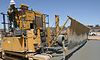A construction worker operates a large yellow paving machine that is spreading and smoothing fresh concrete on a road under a clear blue sky.