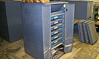 A blue metal storage cabinet with multiple open drawers stands on a concrete floor in an industrial setting. Several flat cases are propped against the wall nearby.