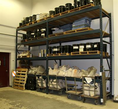 Industrial metal shelving unit with black containers and plastic bins on upper shelves, and bags of materials on carts below. A wooden pallet is leaning against the side.