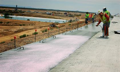 Workers in safety vests and helmets apply curing compound to a concrete surface at a large outdoor construction site with earthmoving equipment in the background.