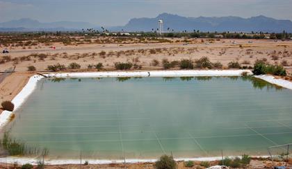 A large, rectangular, man-made pond lined with white material sits in a desert landscape with sparse vegetation and distant mountains under a cloudy sky.