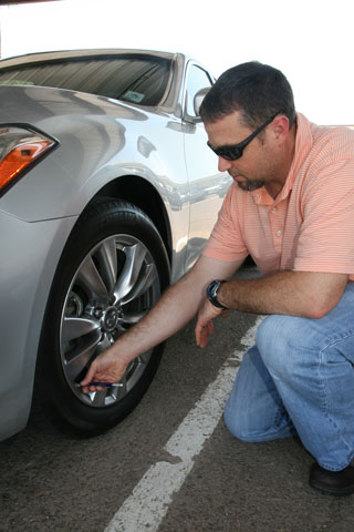 A man wearing sunglasses and a striped polo shirt crouches to check the tire pressure of a parked silver car using a tire pressure gauge.