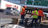 Three construction workers in orange safety vests stand beside a metal guardrail on a highway, with a truck and road signs visible in the background.