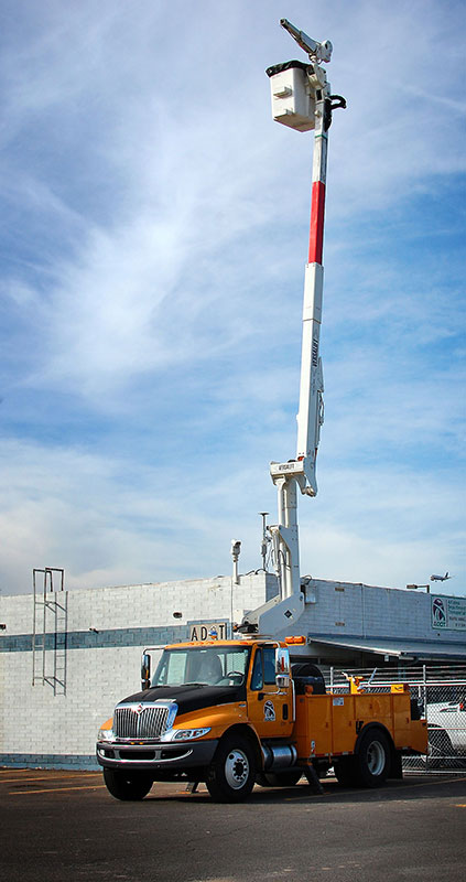 A yellow utility truck with an extended aerial lift holds a worker in a bucket raised high near a building under a partly cloudy sky.