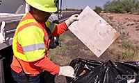 A worker in a safety vest and helmet picks up litter and places it in a black trash bag by the roadside.