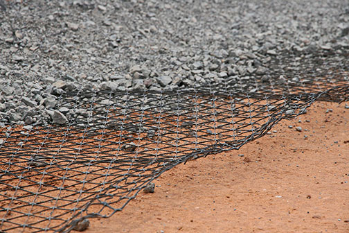 A layer of geogrid mesh lies between compacted gravel above and reddish-brown soil below, illustrating ground reinforcement in construction.