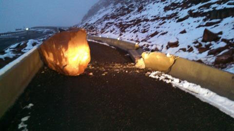 A large boulder blocks a snow-covered mountain road, breaking through a concrete barrier; headlights illuminate the scene in low light.