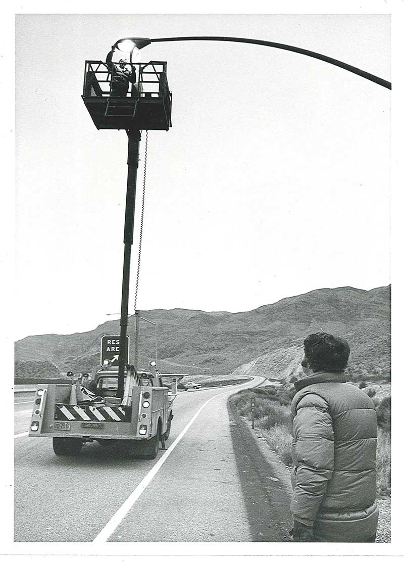 A person stands on the roadside watching a worker in a lift repair a streetlight above a curved road.