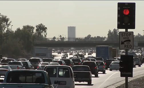 Heavy traffic fills multiple lanes on a freeway under hazy daylight, with a red traffic light and “Stop Here On Red” sign visible in the foreground.