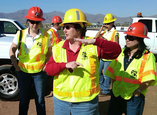 Group of construction workers in hard hats and safety vests talking at a worksite with mountains in the background.