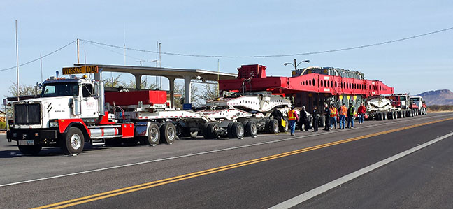 A large truck with an Oversize Load sign hauls an oversized red and white cargo trailer on a highway, accompanied by workers in safety vests.