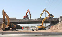 Excavators demolishing a bridge while construction workers oversee the site on a sunny day.
