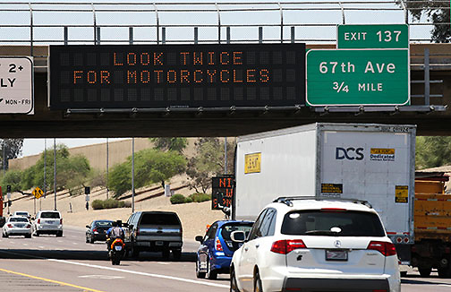 Cars and a motorcycle drive on a highway under a digital sign that reads, LOOK TWICE FOR MOTORCYCLES; exit sign for 67th Ave is visible.