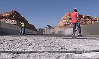Two construction workers in safety gear pour and spread concrete on a bridge, with rocky hills and clear blue sky in the background.
