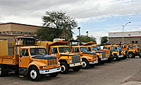 A row of yellow trucks in a parking lot near buildings and trees under a cloudy sky.