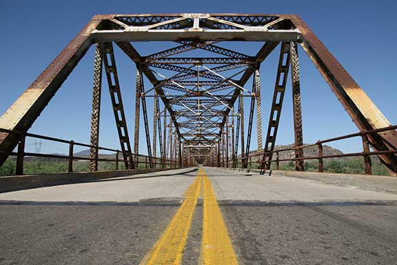 A view down the center of an old, rusted steel truss bridge with a double yellow line on the road, under a clear blue sky.