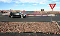 Black minivan enters a roundabout near a yield sign on a clear day with a desert background.