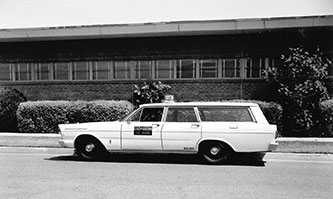 Black and white photo of a vintage station wagon taxi parked in front of a low building with bushes.