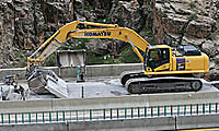 A yellow excavator works beside a concrete barrier on a road in a rocky, mountainous area.