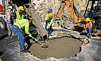 Workers in safety gear pour and spread concrete into a large hole at a construction site with machinery nearby.