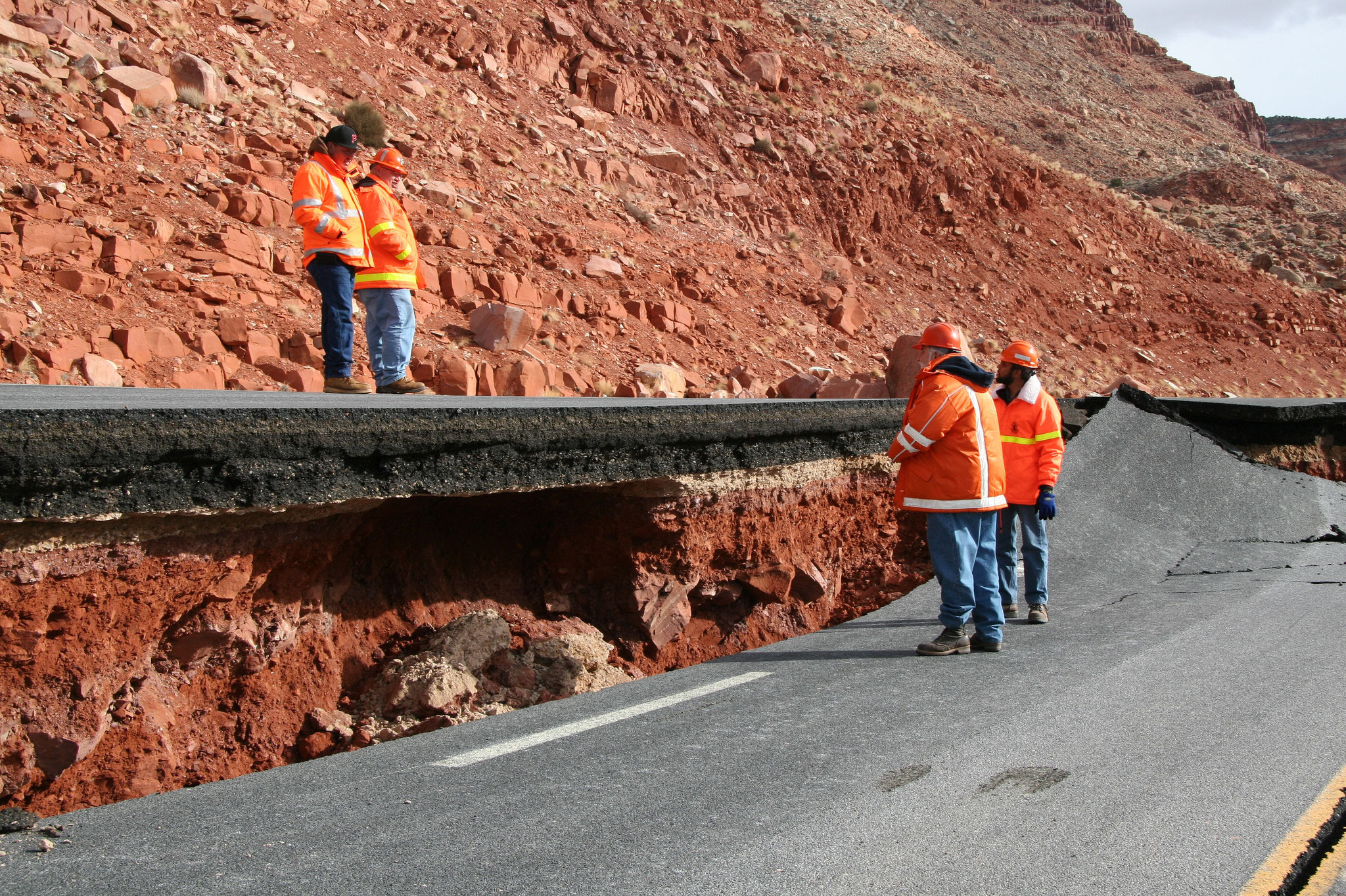 Three workers in orange jackets inspect a road severely damaged and lifted by a landslide in a rocky area.