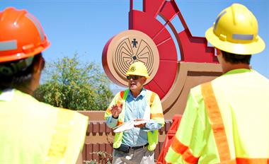 Three construction workers in safety gear talk near a large, red sculptural sign under a clear blue sky.