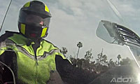 A person wearing a bright yellow and black helmet and safety jacket rides a motorcycle, with trees and a cloudy sky in the background.