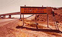 A person sits on a ladder next to a roadside sign reading BLOWING DUST REDUCE SPEED near a highway.