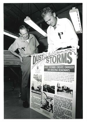 Two men examine a large poster warning about the dangers of dust storms on Arizona roadways.