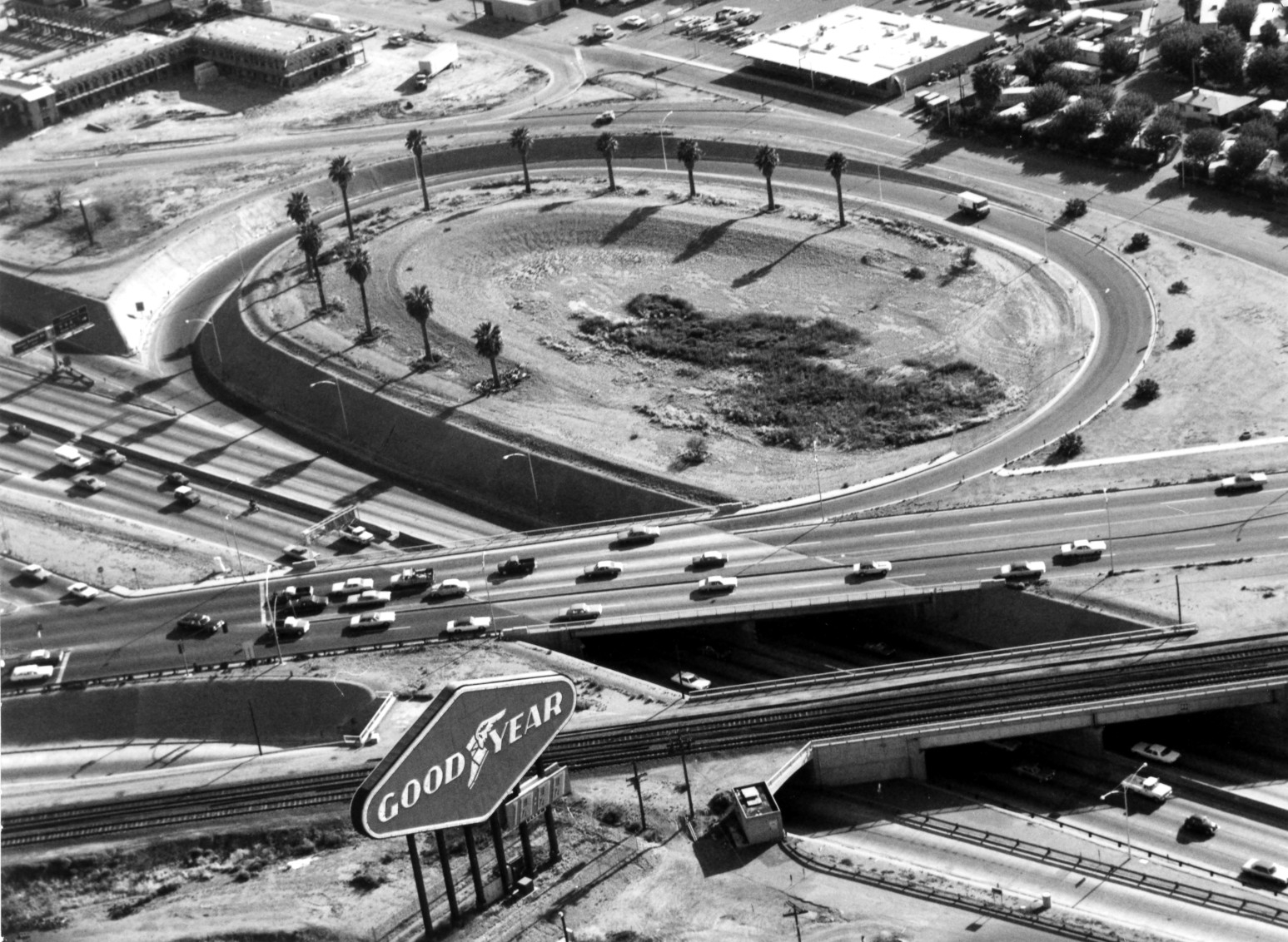 Aerial view of a freeway interchange with multiple cars, a central oval island lined with palm trees, and a large Goodyear sign beside railroad tracks in the foreground.
