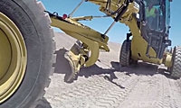A close-up view of a yellow construction grader smoothing a dirt road under a clear blue sky, with visible tire tracks and part of the operator’s cabin.