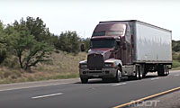 A red semi-truck with a white trailer driving on a highway surrounded by trees and grass.