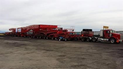 A large red modular trailer with multiple axles is connected to a red semi-truck, parked on a wide gravel lot under an overcast sky.