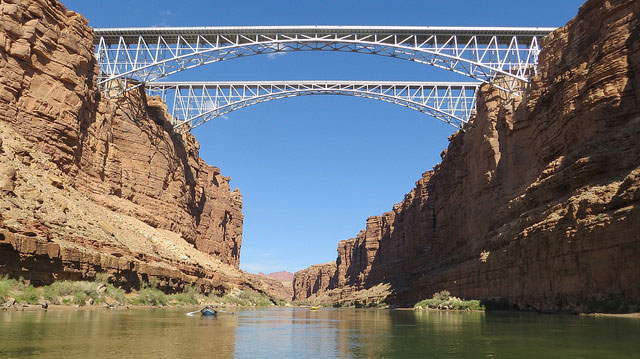 Two steel arch bridges span a deep canyon with red rock walls, above a calm river under a clear blue sky.