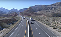 Cars and a truck drive on a divided highway through a desert landscape with mountains in the distance.