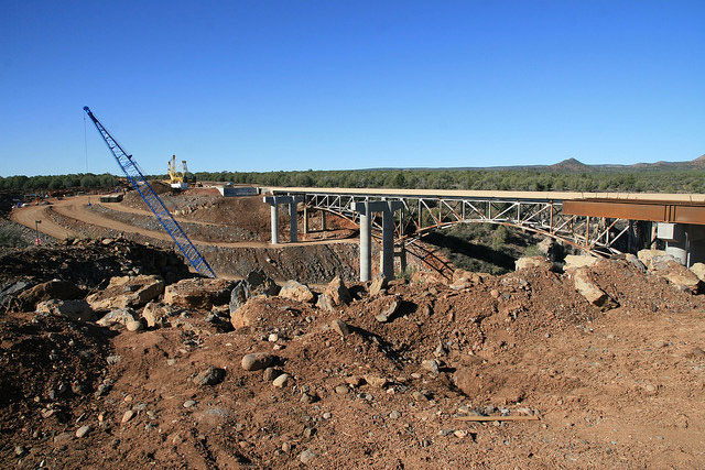 A bridge under construction with a crane, surrounded by dirt and rocks under a clear blue sky.