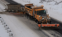 An orange snow plow truck clears snow from a road, with snowy embankments on either side and a salt spreader attached to the back of the truck.