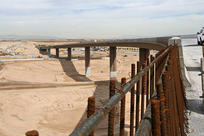 A partially constructed bridge spans a sandy landscape under a cloudy sky, with rusted rebar and parked vehicles visible in the foreground.