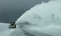 A snowplow clears a snow-covered road, blowing a large plume of snow to the side under a cloudy, gray sky.