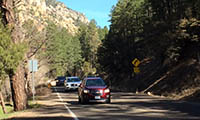 Several cars drive on a winding mountain road surrounded by trees and rocky hillsides under a clear blue sky.