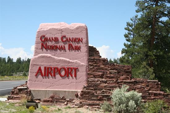 Stone sign reads Grand Canyon National Park Airport with trees, brush, and road in background.