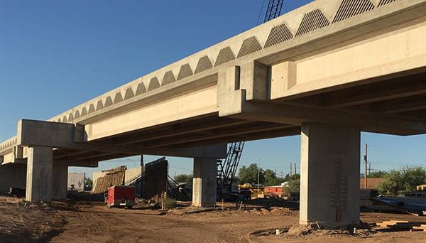 A concrete bridge under construction with support pillars, construction equipment, and dirt visible beneath it on a clear day.